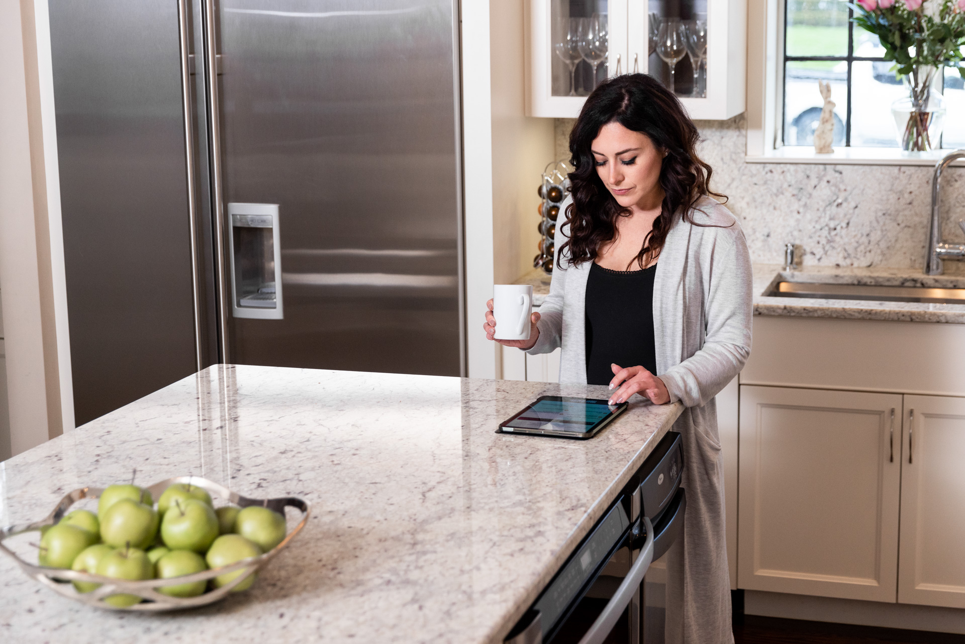 A woman stands at her white marble-topped kitchen island. She is drinking coffee and using a tablet computer.