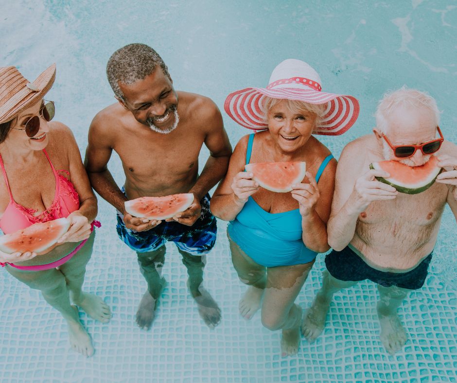 Two older men and two older women stand in a pool, smiling and eating watermelon.