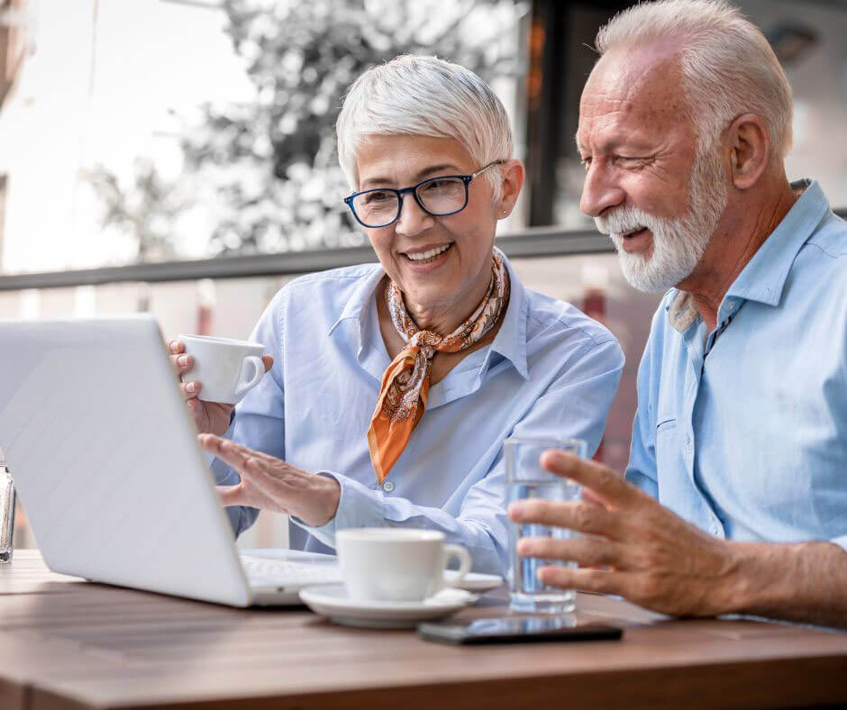 An older couple sits outside and talks to a hearing specialist on their laptop.