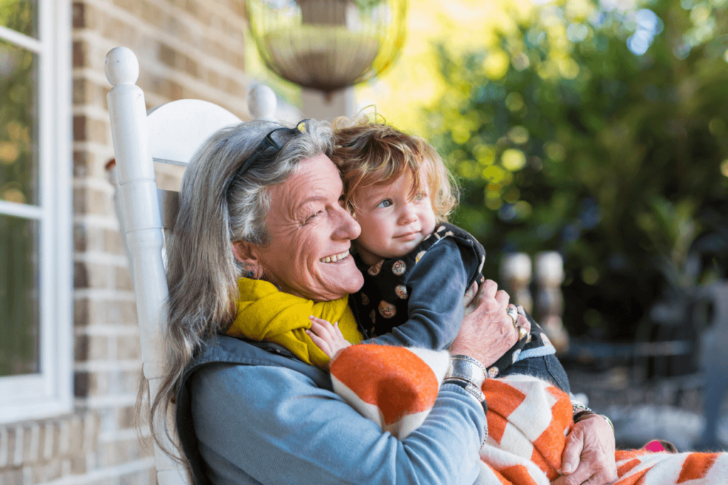 A smiling grandmother holds her toddler grandchild on her lap on a front porch.