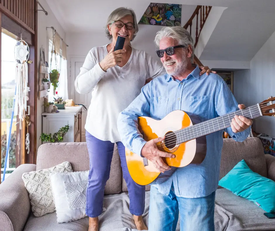 An older woman sings, and an older man plays guitar in their living room.